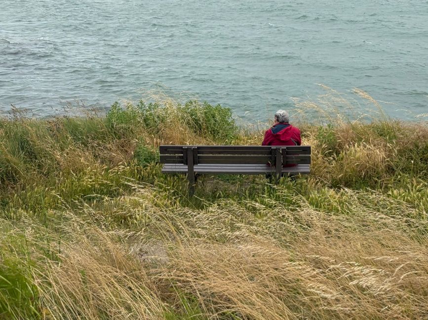 Senior person in red coat sitting alone on a bench by a peaceful lakeside, enjoying nature.