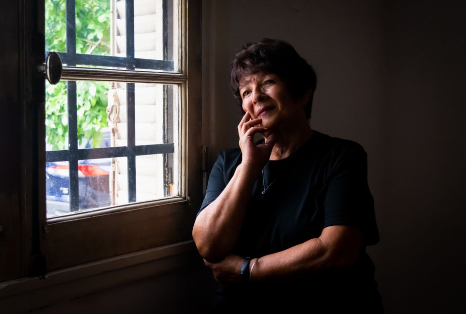A senior woman stands by a window, looking thoughtful and serene in soft, natural light.